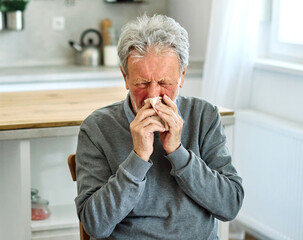 A man is sitting at a table with a tissue in his hand. He is wiping his nose and he is in a state...
