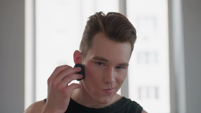 handsome young man in sleeveless signet with neck tattoo sits before bright window mirror, holds black makeup sponge, gracefully dabs powder on cheeks, exudes style and confidence