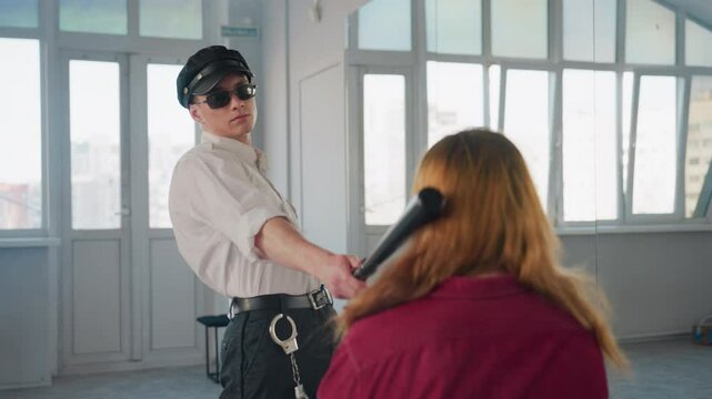 man wearing black cap sunglasses and white shirt grips baton and strikes it on seated woman head while pulling her hair close to face in tense violent act inside bright studio room with windows