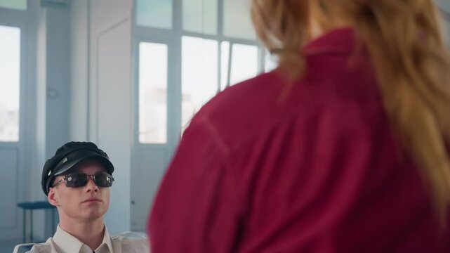rear view of seated woman in red shirt facing male stripper wearing cap sunglasses sliding baton behind head while dancing squatting near mirror in bright minimalist studio with window reflections