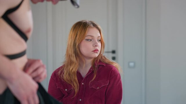 young woman seated in red corduroy shirt watches half dressed male stripper in harness fix trousers then averts gaze before looking back, showing mix of boredom curiosity inside mirrored studio
