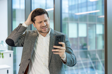 Close-up photo of a shocked and disappointed young businessman standing in the office, holding his...