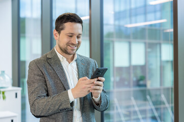 Obraz na płótnie Canvas Close-up photo of a young man in a suit standing smiling in an office building near a window while using a mobile phone