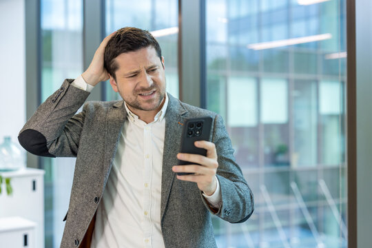 Close-up photo of a shocked and disappointed young businessman standing in the office, holding his head in his hand and looking at the mobile phone screen with concern - Powered by Adobe