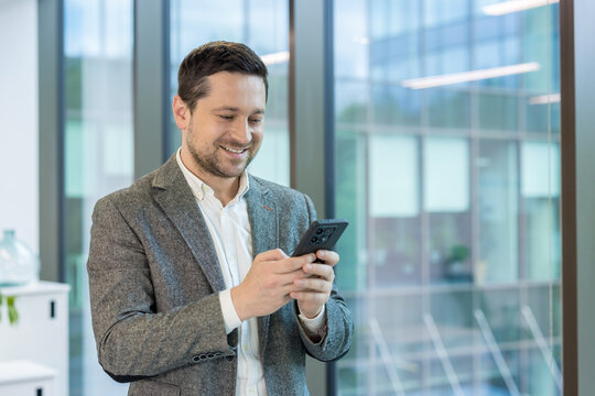 Close-up photo of a young man in a suit standing smiling in an office building near a window while using a mobile phone