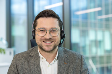 Close-up portrait of a young male office worker sitting at his workplace in a headset and suit and looking at the camera with a smile