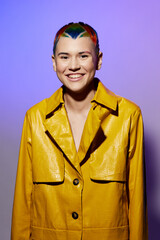 young woman with colorful short hair wearing a yellow jacket smiling confidently against a purple...