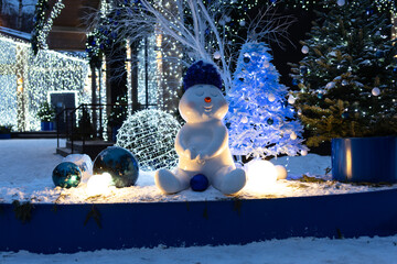 A cheerful snowman stands in a festive urban setting. Decorated with elegant Christmas trees and twinkling lights, creating the atmosphere of a winter holiday.