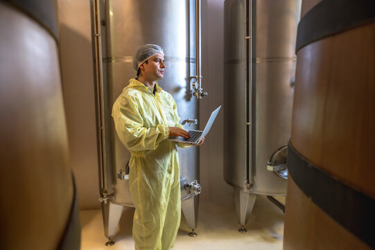 Expertise modern male wine cellar  maker holding laptop checking quality control and creating test trail in stainless steel barrels 