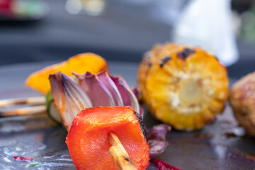 Plate of food with a skewer of vegetables and a piece of corn. The vegetables and the corn are grilled.