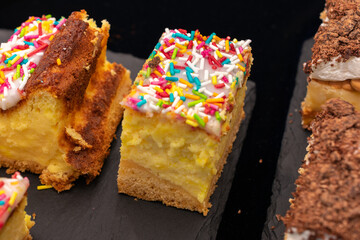 Plate of assorted desserts with colorful sprinkles on top. The desserts are arranged in a way that they look like they are cut into small pieces. The plate is placed on a black surface.