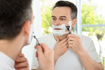 Handsome man shaving with razor near mirror at home