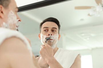 Handsome man shaving with razor near mirror at home, low angle view