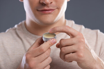 Young man applying lip balm onto his lips on grey background, closeup