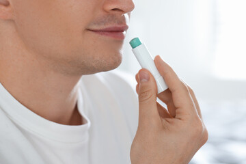 Young man applying lip balm onto his lips at home, closeup