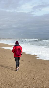 Slow motion 4K vertical footage female with red backpack walking by wide sandy beach at Aguda settlement near Porto, Portugal. Environment, travelling, recreation concept.