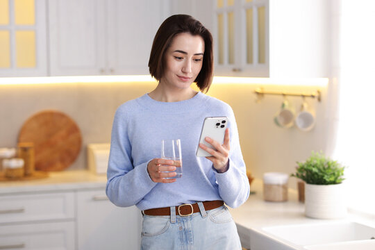 Woman with glass of water using modern smartphone in kitchen