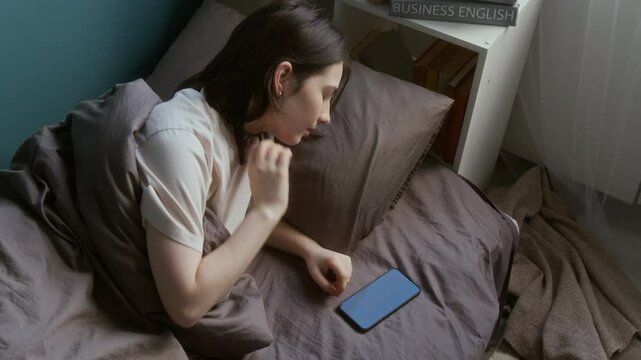 High angle shot of brunette teenage girl waking up in morning and picking up smartphone texting while lying in bed