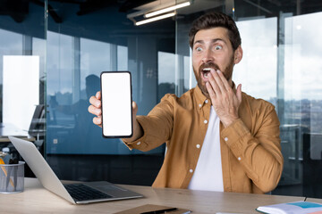 A man in an office is surprised while holding a blank smartphone screen, ideal for showcasing app...