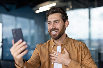 A smiling man holds a phone, likely during a video call, expressing joy and friendliness in an...