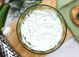 Making Tzatziki Greek or Mediterranean sauce or dip for a party. Dip or sauce in a green bowl on a wooden cutting board with dill, green napkin and a grater.  Greek Tzatziki sauce for all foods.