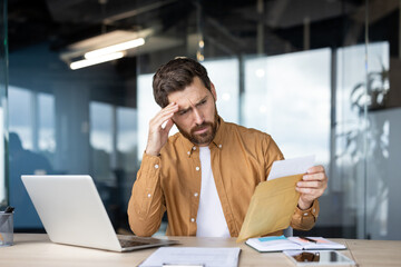 A concerned businessman reviews documents while working at his desk, appearing stressed and...