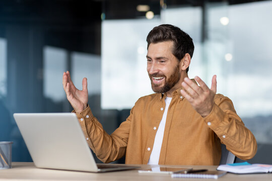 A businessman is happy and animated, with his hands raised in excitement, while participating in a video call using a laptop.