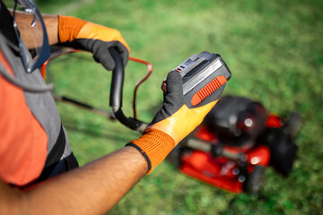 Landscape maintenance process with man holding a battery against lawn mower in a garden setting during daylight hours
