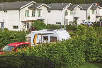 White travel trailer and red car parked by trimmed green hedge near residential houses after summer rain. Sweden.