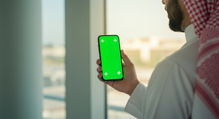 Arab businessman holding smartphone with green screen, near window.