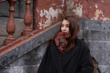 Woman in a cozy coat gazing thoughtfully while standing against a weathered wall during a chilly...