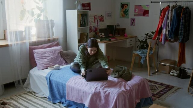 Wide angle shot of young teenage girl watching TV show via laptop and laughing while sitting on bed in colorful dorm room