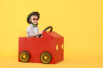 Little boy driving car made of cardboard on yellow background. Space for text