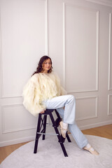 A portrait of a fashionable teenage girl in blouse and sneakers sitting on a chair in a studio on a light wall in a room. light from the window
