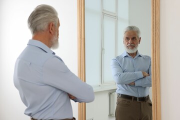 Handsome senior man looking at himself in mirror indoors