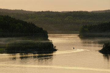A boat cruises through a peaceful, mist-covered lake surrounded by lush green forests at sunset,...