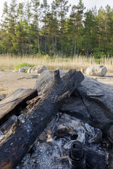 Burnt wood and ashes from a previous fire lie on a rocky lakeside, with a backdrop of tall trees and a peaceful shore.