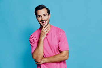 Smiling happy cheerful young man wearing pink t-shirt looking at camera with hand on chin isolated...