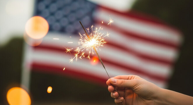 Sparkler in hand with blurry U.S. flag background.
Hand holding glowing sparkler in front of large American flag, festive and warm mood.