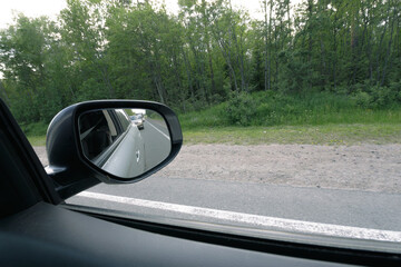 View from a car window showing vehicles behind in the side mirror, with a green forest lining the...