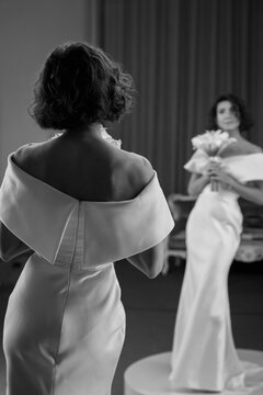 Stylish bride in an off-shoulder gown is seen from behind, admiring her reflection in the mirror in a timeless black and white shot. Elegant Bride Reflected in Mirror