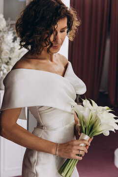 Happy bride in an off-shoulder white gown poses with a bouquet in a stylish, softly lit dressing room. Smiling Bride Holding Flowers in Elegant Dressing Room