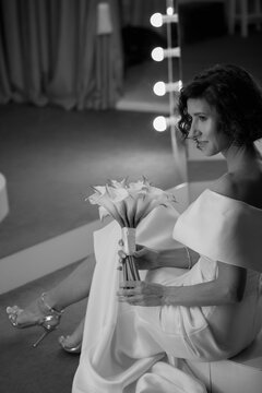 Bride in a white off-shoulder gown sits gracefully near a lit vanity mirror, holding flowers in a stylish dressing room. Elegant Bride Sitting by Mirror in Glamorous Dressing Room