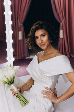 Happy bride in an off-shoulder white gown poses with a bouquet in a stylish, softly lit dressing room. Smiling Bride Holding Flowers in Elegant Dressing Room