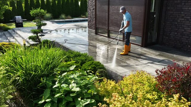 Man Cleaning Patio With Pressure Washer in Residential Backyard During Sunny Day