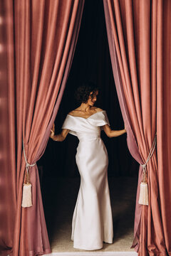 Stylish bride in a fitted off-shoulder gown posing gracefully between parted pink velvet curtains in a dramatic setting. Elegant Bride Framed by Pink Velvet Curtains