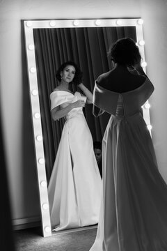 Stylish bride in an off-shoulder gown is seen from behind, admiring her reflection in the mirror in a timeless black and white shot. Elegant Bride Reflected in Mirror
