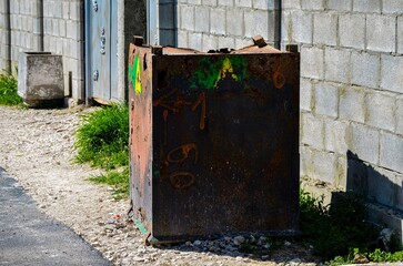 A rusted metal box with graffiti on it sits on the side of a road. The box is covered in green and yellow paint, and the graffiti includes the letters A and B. The box is surrounded by grass and weeds