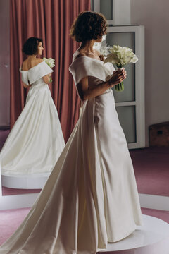 Happy bride in an off-shoulder white gown poses with a bouquet in a stylish, softly lit dressing room. Smiling Bride Holding Flowers in Elegant Dressing Room