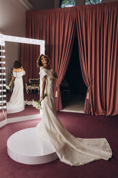 Happy bride in an off-shoulder white gown poses with a bouquet in a stylish, softly lit dressing room. Smiling Bride Holding Flowers in Elegant Dressing Room